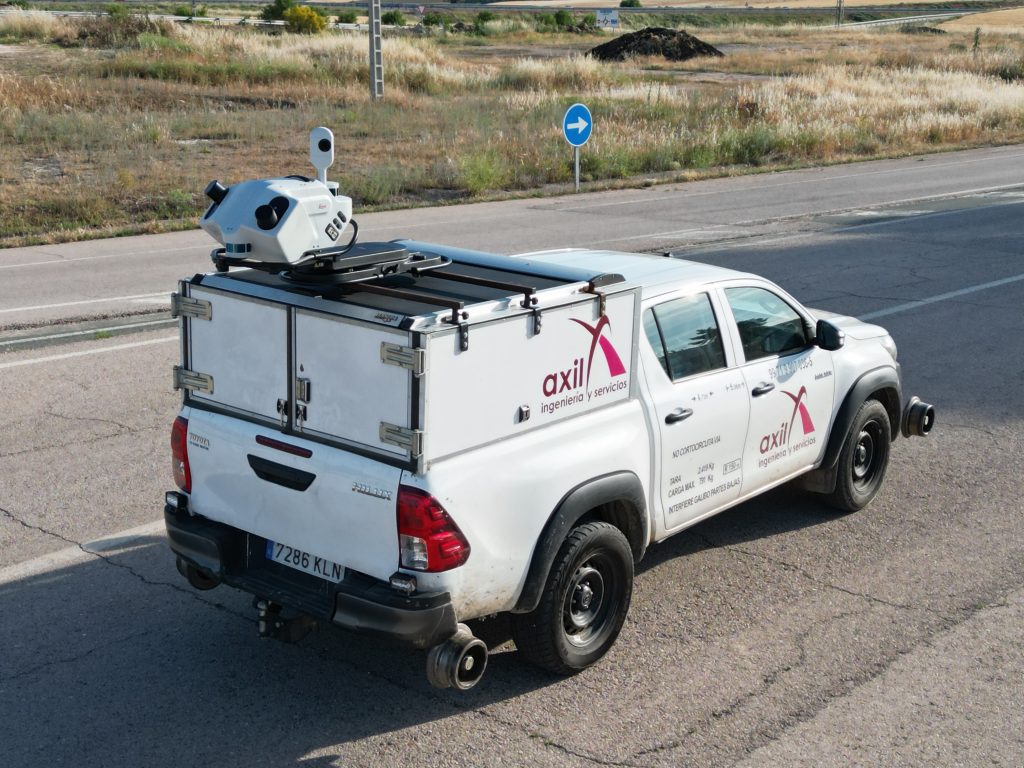 White service van with rooftop surveying equipment mounted on top, parked along a road with dry field behind it.